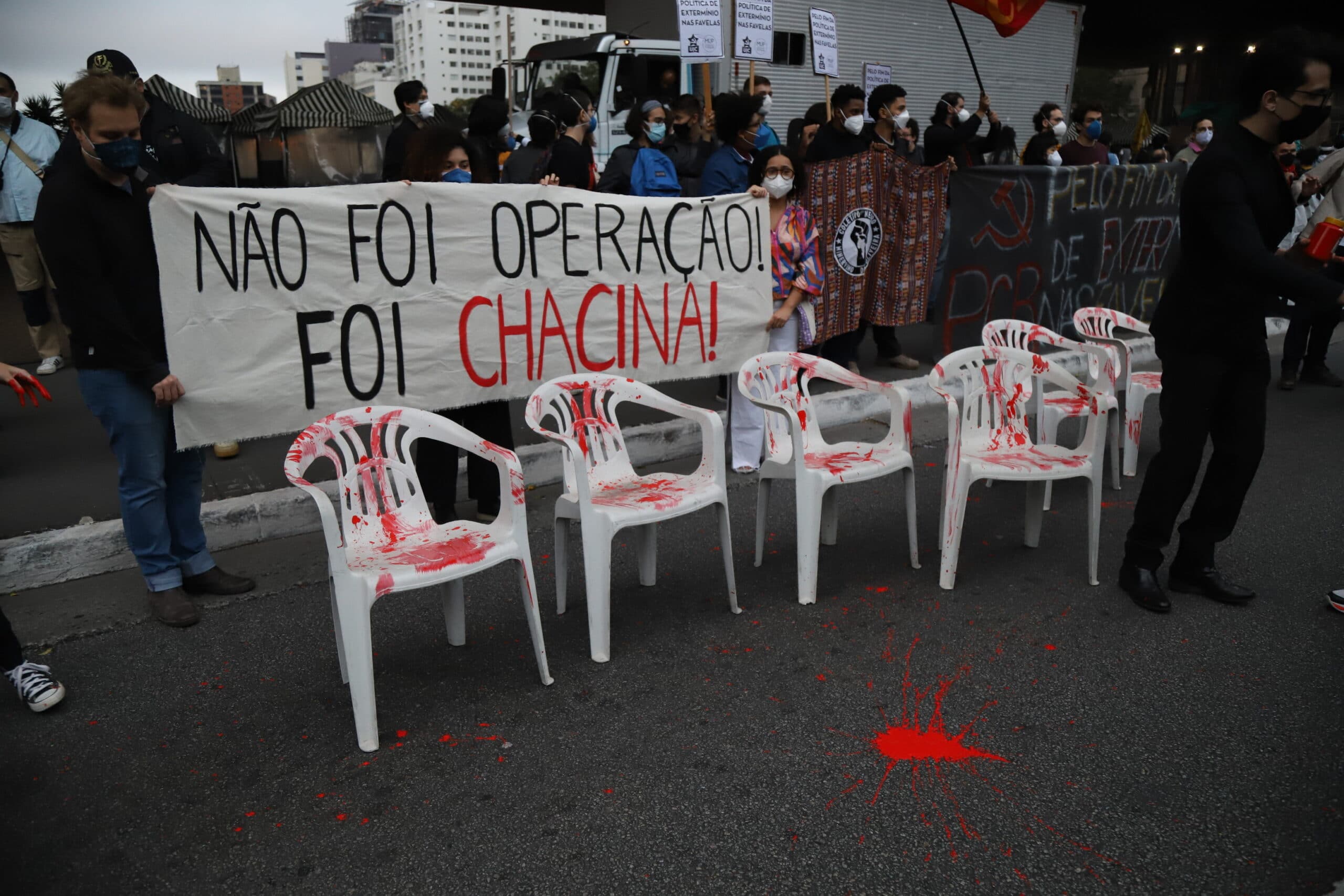 Manifestantes pintam de vermelho quatro cadeiras simulando sangue. Atrás, uma faixa diz 